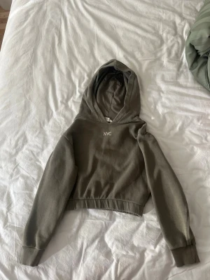 Olivgrön hoodie med NYC-tryck - Snygg olivgrön hoodie med broderat NYC-tryck på bröstet. Croppad modell med långa ärmar, ribbade muddar och huva. Perfekt för en avslappnad och trendig stil. Mjuk bomullskänsla och enkel att matcha med jeans eller leggings.