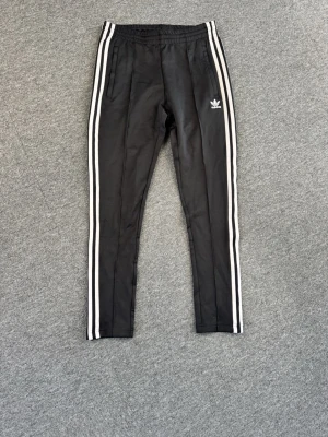 Svarta Adidas mjukisbyxor med vita ränder - Säljer ett par svarta mjukisbyxor från Adidas med klassiska vita ränder längs benen och Adidas-logga på vänster sida. Byxorna har smal passform, resår i midjan och är tillverkade i ett mjukt och bekvämt material. Perfekta för chill.