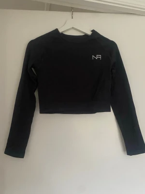 Svart croppad långärmad träningstopp NA - Svart croppad långärmad tränings topp från NA med enkel design och liten vit logga på bröstet. Perfekt för en sportig och trendig look. Mjuk och stretchig känsla, sitter tight mot kroppen.
