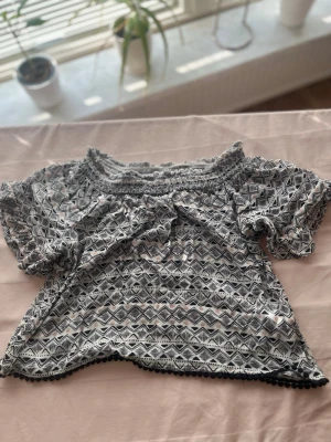 Svartvit offshoulder blus med mönster - Säljer en luftig svartvit offshoulder blus med grafiskt mönster och små bollar längs nederkanten. Blusen har korta ärmar med resår och är gjord i ett tunt, mjukt tyg som känns skönt mot huden. Perfekt till jeans eller kjol för en chill look. Andvänt 1 gång!