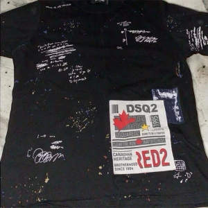 Svart DSQUARED2 t-shirt med tryck - Cool svart t-shirt från DSQUARED2 med unikt tryck, färgstänk och grafiska detaljer framtill. T-shirten har korta ärmar och flera vita texttryck samt en stor patch med röd och vit design. Perfekt för dig som gillar streetstyle.