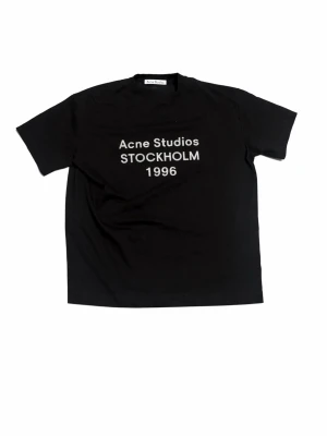 Svart Acne Studios t-shirt - Svart t-shirt från Acne Studios med vit text 'Acne Studios STOCKHOLM 1996' på bröstet. Klassisk passform och rund halsringning. Perfekt för dig som gillar stilrena och trendiga plagg med tydlig logga.