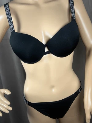 Svart set från Victoria's Secret - Snyggt svart underklädesset från Victoria's Secret med vadderad bh och matchande trosa. Axelbanden har logotypdetaljer i vitt och silver. Setet har en modern och sportig look med mjukt material och diskret cut-out framtill på bh:n.