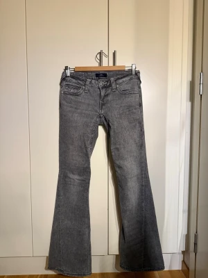 Grå bootcut jeans från True Religion - Säljer ett par gråa bootcut jeans från True Religion med klassiska kontrastsömmar och fem fickor. Jeansen har låg midja, utsvängda ben och detaljerade bakfickor med knappar. Perfekta för en avslappnad men trendig look.