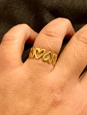 Stål Guldfärgad ring med hjärtan - Säljer en söt guldfärgad ring med öppna hjärtan runt hela bandet. Ringen har en modern och lekfull design som sticker ut och passar perfekt för dig som gillar detaljer. Materialet är metall med en blank finish. Perfekt accessoar för att lyfta din stil.
