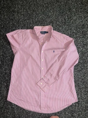 Rosa randig skjorta från Ralph Lauren - Klassisk rosa och vit randig skjorta från Ralph Lauren med button-down krage och lång ärm. Skjortan har broderad blå logga på bröstet och vita knappar framtill. Tillverkad i bomull för en skön och luftig känsla. Väldigt fin skjorta inför sommaren. Använd fåtal gånger och är i väldigt fint skick. Tveka inte på att höra dig vid intresse. Pris kan alltid diskuteras/Isak😀