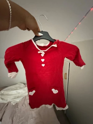 Röd tomteklänning med hjärtan - Söt röd tomteklänning med vita detaljer, hjärtformade knappar och små hjärtan på fickorna. Klänningen har långa ärmar och vita muddar vid ärmslut och nederkant. Perfekt för julens maskerader och temafester.