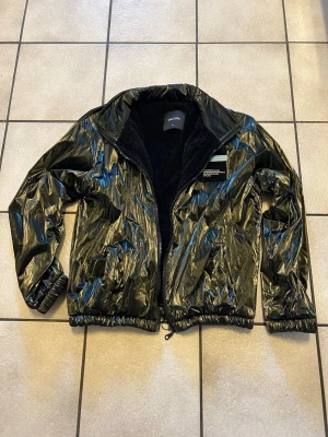 Svart glansig vindjacka från qui life - Cool svart vindjacka från qui life med glansig finish och dragkedja framtill. Jackan har en mjuk, svart fleecefodrad insida och resår vid ärmslut och midja. Detaljerad med patch och text på bröstet för en modern look.