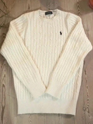Vit kabelstickad tröja från Polo Ralph Lauren - Kabelstickad vit tröja från Polo Ralph Lauren med rund halsringning och diskret broderad logga på bröstet. Tröjan har långa ärmar och ribbade muddar vid ärmslut och nederkant. Perfekt för lager på lager och klassisk stil. Har påsen kvar 