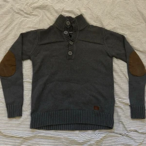 Grå stickad tröja Jack & Jones - Mörkgrå stickad tröja från Jack & Jones med hög krage och fyra stora knappar framtill💫 Tröjan har ribbade muddar och nederkant samt bruna mockadetaljer på armbågarna. Perfekt för en snygg och avslappnad stil🍀