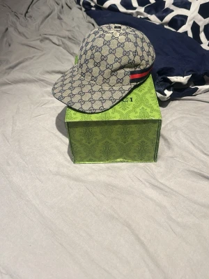 Gucci keps med GG-mönster - Snygg keps från Gucci i beige canvas med klassiskt GG-monogram över hela kepsen. Har en böjd skärm och en röd-grön rand på sidan som ger en lyxig touch. Perfekt accessoar för att lyfta din outfit och visa din stilkänsla.