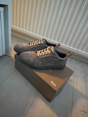 Gråa sneakers med röd sula Paris - Stilrena gråa sneakers i mocka med klassisk låg profil och röda sulor. Skorna har beigea skosnören och diskreta sömmar för en clean look. Perfekta för dig som gillar minimalistisk design och vill sticka ut med en färgglad detalj under foten.