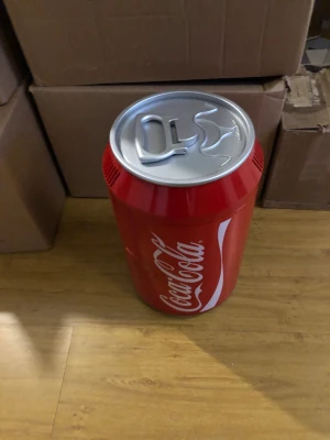 Coca-Cola minikyl i burkdesign - Säljer en röd minikyl i form av en Coca-Cola burk. Kylen har ett silverfärgat lock, vit Coca-Cola-logga och ett hyllplan inuti. Perfekt för att förvara drycker eller snacks. Tillverkad i plast och metall.