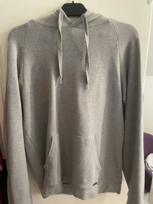 Grå Boss hoodie med ficka - En stilren grå hoodie med huva och dragsnören. Tröjan har en klassisk känguruficka framtill och långa ärmar. Perfekt för en avslappnad look. Materialet är mjukt och bekvämt, troligtvis bomullsblandning. Pris kan diskuteras 