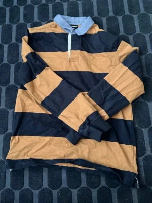 Randig rugbytröja med krage - Snygg långärmad rugbytröja från H&M med breda svarta och bruna ränder samt klassisk grå krage. Tröjan har normal passform och är tillverkad i bomull, perfekt för en avslappnad stil.