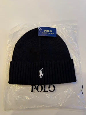 Svart mössa/beanie från Polo Ralph Lauren - Snygg svart ribbstickad mössa från Polo Ralph Lauren med klassisk broderad logga framtill. Mössan har uppvikt kant och är tillverkad i mjukt material som håller dig varm. Perfekt accessoar för kyliga dagar och stilren look. Snabb frakt. Skickas samma dag, helt oanvänd. Hör av dig vid frågor eller annat.🤗😁