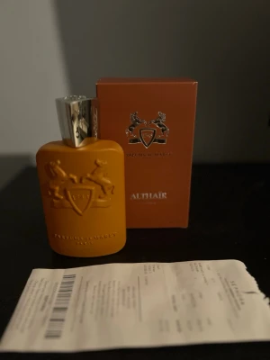 Parfums de Marly Althaïr parfym - Exklusiv parfym från Parfums de Marly, modellen Althaïr. Flaskan är matt orange med silverfärgad kork och har ett elegant reliefmönster med logotyp. Kommer i matchande orange kartong med stilren design och detaljerad text på baksidan.