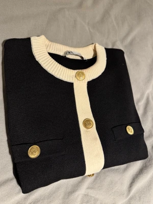 Svart kofta med gulddetaljer - Svart tröja med krämvita ribbade kanter och dekorativa guldfärgade knappar framtill och på fickorna. Rund halsringning och klassisk design som passar till mycket. Materialet är stickat och känns mjukt. Använt 1 gång, då jag fick smink på armen som syns på bilden men kan nog enkelt tvättas bort.