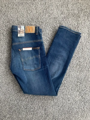 Blå jeans från Nudie Jeans - Säljer ett par klassiska blå jeans från Nudie Jeans med raka ben och snygga slitningar. Jeansen har fem fickor, orange sömmar och patch med logga bak i midjan. Tillverkade i bomull med en cool tvättad look och diskreta detaljer.