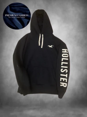 Svart hoodie från Hollister - Svart hoodie från Hollister med vit logga på bröstet och stort vitt tryck längs ena ärmen. Klassisk känguruficka framtill och vita dragsnören i huvan. Perfekt för en avslappnad och trendig stil.