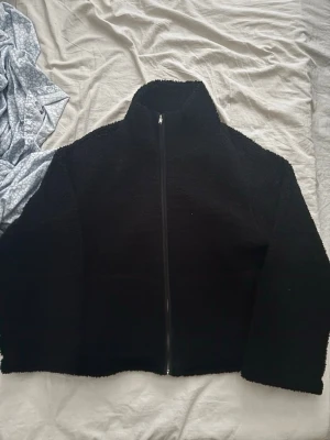 Svart teddyjacka från 157 - Mysig svart teddyjacka från 157 med hög krage och hel dragkedja framtill. Jackan har en fluffig och mjuk textur som ger en avslappnad vibe. Perfekt för att hålla värmen under kyliga dagar och enkel att matcha med olika outfits.