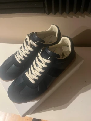 Maison margiela skor  - Snygga mörkblå sneakers med vita snören och beige innersula. Skorna har en klassisk låg siluett och detaljer i mocka och textil. Yttersulan är i gummi med naturfärg. Perfekta för dig som gillar stilrena och tidlösa sneakers.