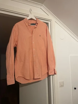 Persika/rosa färgad skjorta från Ralph Lauren - Säljer en persika/rosa färgad skjorta från Ralph Lauren med klassisk button-down krage och broderad logga på bröstet. Skjortan har långa ärmar och är tillverkad i mjuk bomull. Perfekt för en avslappnad men ändå stilren look.