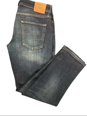 Mörkblå jeans från Jack & Jones taperd mike  - Snygga mörkblå jeans från Jack & Jones med klassisk femficksdesign och diskreta slitningar. Jeansen har raka ben och en läderpatch med logga bak i midjan. Tillverkade i bomull med en skön passform som funkar till många stilar. Hör av er via funderingar 
