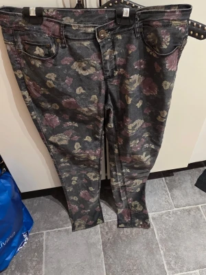 Blommiga skinny jeans  - Säljer ett par skinny jeans med blommigt mönster i beige och vinröda toner. Jeansen har klassisk femficksmodell, knappgylf och är tillverkade i stretchigt denimtyg för skön passform. Perfekta för dig som vill sticka ut med en cool och unik stil.