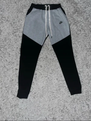 Nike grå och svarta mjukisbyxor - Snygga mjukisbyxor från Nike i grått och svart med dragsko i midjan och fickor på sidorna. Byxorna har en sportig look med kontrastpaneler och Nike-logga på låret. Perfekta för chill eller träning.platsen där dragsnörena sitter är skadad. Size Xl men passar L. Priset diskutera.
