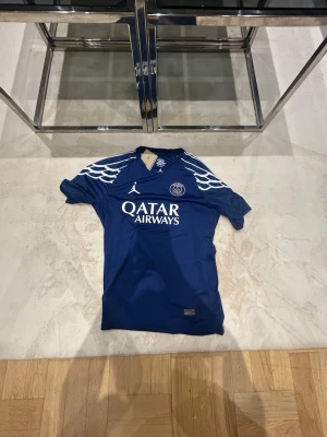 PSG matchtröja blå Jordan x Qatar - Snygg blå PSG matchtröja med vita detaljer, Jordan-logga och Qatar Airways-tryck på bröstet. Nummer 14 och D. Doué på ryggen samt Champions League-märke på ärmen. Perfekt för fotbollsfans som vill sticka ut på läktaren eller planen.