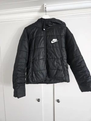 Svart pufferjacka från Nike - Snygg svart pufferjacka från Nike med huva och dragkedja. Jackan har vit Nike-logga på bröstet och en stort nike märke på ryggen. Perfekt för kalla dagar och streetwear-stil. Storlek s