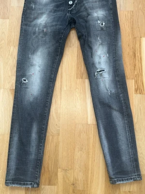 Grå slitna jeans med Pac-Man-knappar - Säljer ett par grå jeans med slitningar och distressed detaljer samt coola Pac-Man-inspirerade knappar i gylfen. Jeansen har en tvättad look med ljusa partier och små färgstänk. Perfekta för dig som gillar en unik och trendig stil.