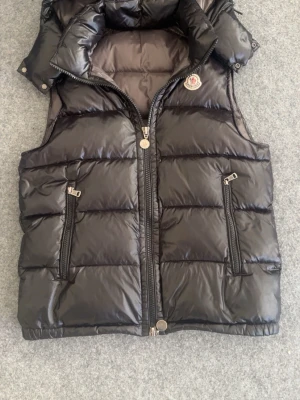 Svart dunväst från Moncler - Snygg svart dunväst från Moncler med hög krage och två dragkedjeförsedda fickor framtill. Västen har quiltad design och klassisk Moncler-logga på bröstet. Insidan har ett unikt serietidningsmönster och märkestaggar. Perfekt för lager på lager och streetwear.
