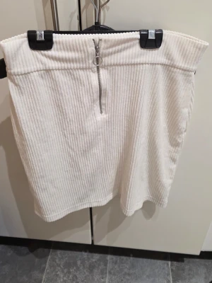 Vit ribbad manchesterkjol med dragkedja - Säljer en vit kortkjol i ribbad manchester med en snygg silverfärgad dragkedja framtill och ringdetalj. Kjolen har en hög midja och är perfekt för dig som gillar en clean och trendig stil.