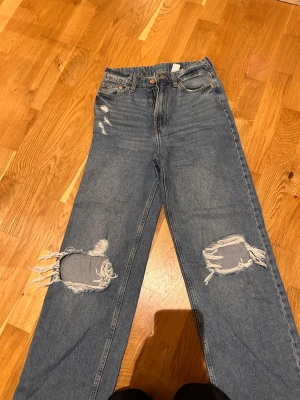 Blå slitna mom jeans med hål - Säljer ett par blå mom jeans med hög midja och stora slitningar på båda knäna. Jeansen har raka ben, klassisk femficksmodell och råa kanter vid bensluten. Perfekta för en avslappnad och trendig look.