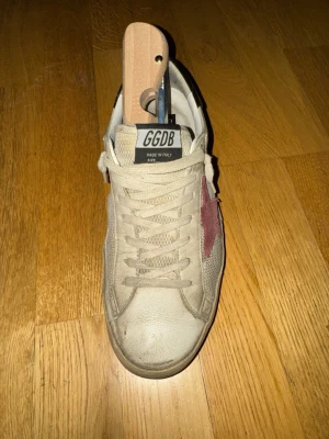 Golden Goose - Snygga beige sneakers från Golden Goose med en ikonisk röd stjärna på sidan och svart detalj vid hälen. Skorna har vita snören och är tillverkade i en mix av läder och textil. Perfekta för dig som gillar en avslappnad men trendig stil. Dem är ganska slitna med små hål vid hällarna (hör av dig om du har några frågor eller vill ha mer bilder på skorna)