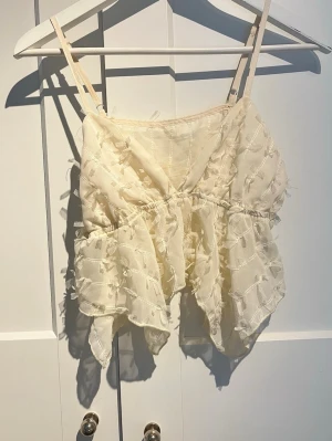 Krämvit volangig topp med tunna band - Söt krämvit topp med tunna axelband och volangig nederdel. Toppen har ett transparent, strukturerat tyg med små dekorativa detaljer och justerbara band. Perfekt för en trendig och luftig look i sommar.