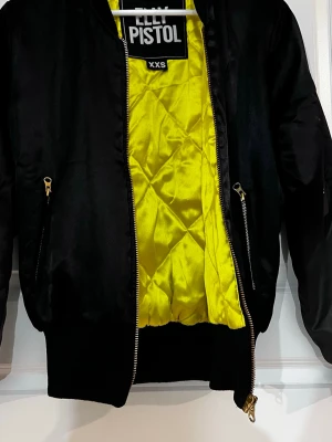 Svart bomberjacka från Elly Pistol - Snygg svart bomberjacka från Elly Pistol med neongul quiltad insida och guldfärgade dragkedjor. Jackan har ribbade muddar vid ärmslut och midja samt fickor med dragkedja och en ärmficka med logotyp. Materialet är glansigt och ger en cool streetwear-look. Passar även upp till small. Mindre slitage på insidan på fodret, se bild.