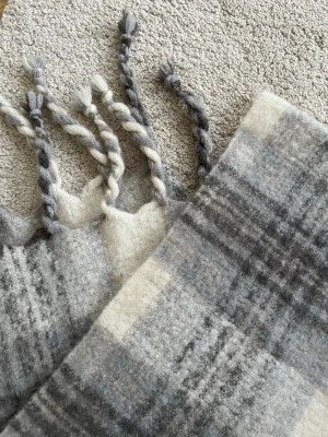 Gråmelerad ullhalsduk med tofsar - Mysig halsduk i mjuk ull med gråmelerat rutmönster och tvinnade tofsar i ändarna. Färgskalan går i olika nyanser av grått och vitt, vilket ger en stilren och klassisk look. Perfekt för att hålla värmen under kyliga dagar.