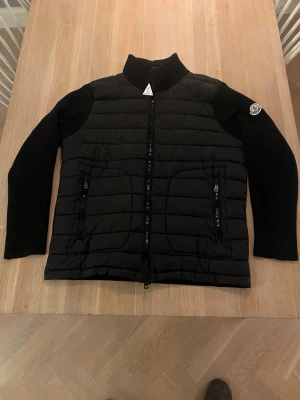 Svart cardiganjacka från Moncler - Svart cardiganjacka från Moncler med quiltad front och ribbstickade ärmar och rygg. Jackan har två dragkedjefickor fram, hög krage och Moncler-logga på ena ärmen. Perfekt för lager-på-lager och snygg streetstil.