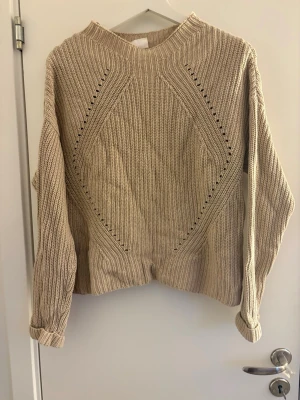 Beige stickad tröja från Stories - Snygg beige stickad tröja från Stories i storlek S (36-38). Tröjan har ett ribbstickat mönster med dekorativa håldetaljer framtill och rund halsringning. Perfekt för lager på lager och enkel att matcha med jeans eller kjol.