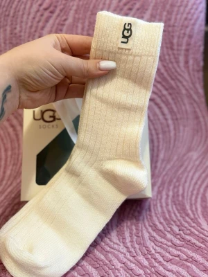 Vita ribbade strumpor från UGG - Säljer ett par vita ribbade strumpor från UGG. De är höga, har en klassisk UGG-logga upptill och är tillverkade i mjukt material som känns riktigt skönt mot huden. Perfekta för dig som vill ha både stil och komfort.