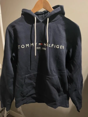 Mörkblå hoodie från Tommy Hilfiger, st. M - Snygg mörkblå hoodie från Tommy Hilfiger med vit broderad logga och text på bröstet. Storlek M