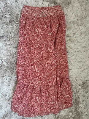 Röd paisleymönstrad långkjol Zara - Boho-inspirerad långkjol från Zara i rött och vitt med paisleymönster och smock i midjan. Kjolen har en avslappnad passform och är tillverkad i ett mjukt, lätt material som passar perfekt till varmare dagar.