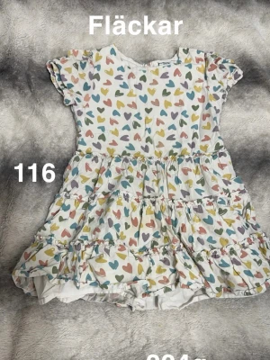 Vit klänning med färgglada hjärtan - Söt vit klänning med kort ärm och volangkjol, täckt av färgglada hjärtan i blått, rosa, gult och grönt. Klänningen har rund hals och lekfull design, perfekt för sommarens alla tillfällen.🌸 Fläckar. Storlek 116