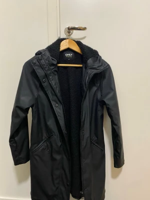 Svart regnjacka från ONLY med foder - Snygg svart regnjacka från ONLY med mysigt teddyfoder på insidan. Jackan har huva, dragkedja och tryckknappar framtill samt två fickor med lock. Perfekt för regniga dagar och håller dig både torr och varm.