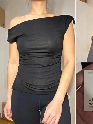 Svart asymmetrisk topp Afternoon - Snygg svart topp med asymmetrisk axel från Afternoon in the House. Toppen har rynkade detaljer i sidan och en liten slits nedtill. Tillverkad i stretchigt material som sitter tajt och bekvämt. Perfekt för en trendig och modern look.