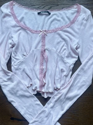 Ljusrosa topp med spets och rosett - Supersöt ljusrosa topp med långa ärmar, spetsdetaljer längs ringningen och framtill samt en liten rosett vid halsen. 