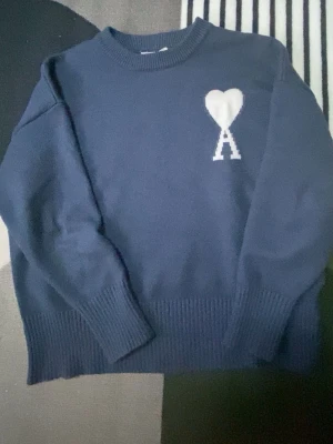 Mörkblå stickad tröja från Ami Paris - Mörkblå stickad tröja från Ami Paris med deras ikoniska vita hjärta och A-logga på bröstet. Tröjan har rund hals, ribbstickade muddar och är gjord i ett mjukt material. Perfekt för en stilren och trendig look.